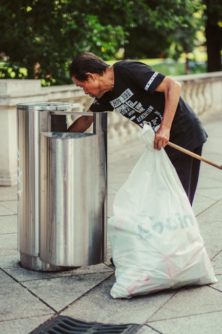A middle-aged man wearing a black T-shirt with white printed text and dark trousers is seen discarding rubbish into a tall, stainless steel, dual-compartment litter bin on a paved outdoor walkway. The man is bent forward, using his right hand to place a white plastic bag filled with waste into the bin, which is positioned beside a low concrete wall with decorative balustrades. The surrounding environment features lush green trees and well-maintained greenery in the background, suggesting a park or garden setting. The scene is illuminated by natural daylight, highlighting the metallic finish of the waste receptacle and the casual attire of the individual. This image visually emphasizes the importance of proper waste disposal, relevant to independent rubbish collection and on-site clearance services provided by [COMPANY_NAME], demonstrating responsible rubbish handling in public outdoor spaces.