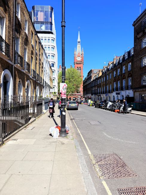 A wide urban street scene in daytime with a clear blue sky, featuring a row of brown brick and stone residential buildings with arched windows and black iron railings along the sidewalk on the left. The concrete pavement is clean, with a few pedestrians walking, some near a white plastic bag placed on the sidewalk. A black lamppost stands centrally, with a traffic sign attached. To the right, parked motorcycles and cars line the street near the curb. In the background, a prominent red and white clock tower with a pointed spire rises above the surrounding buildings, with a mix of modern and historic architecture visible. The scene portrays a typical city environment, with a focus on the street's layout and building facades, reflecting urban residential and commercial areas, and illustrating alternative waste removal convenience through curbside rubbish placement, relevant to private waste disposal services like those offered by Rubbish Collection Earls Court.