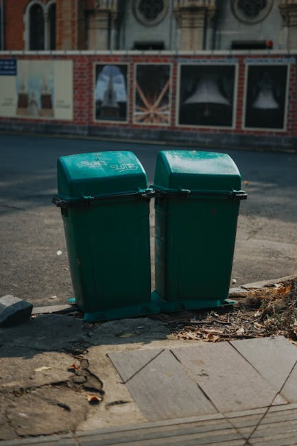 Two green wheelie bins with hinged lids, made of plastic with a slightly textured surface, are positioned side by side on the pavement at the edge of a street. The bins are marked with white text indicating they are for waste collection and are fitted with black handles securing the lids. They sit on a concrete surface that shows some dirt and debris around the base, with a few small leaves scattered nearby. Behind the bins, part of a weathered brick wall is visible, with large posters or advertisements mounted on it, slightly obscured and faded. The background includes a traffic road with grey asphalt, and a building façade with ornate architectural details and windows. The scene is lit by natural daylight, providing clear visibility of the bins and surrounding environment, typical of an urban street setting where private rubbish collection may be arranged through local waste management services like those offered by rubbishcollectionearlscourt.co.uk for alternative rubbish removal options in the area.