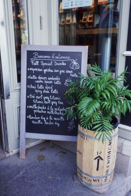 A black chalkboard menu is positioned on a wooden A-frame stand outside a commercial establishment, listing various fall special drinks such as espresso geisha san jose, latte épices amberes, warm spices latte, chai latte épice, and others. The menu is handwritten in white chalk with simple decorative elements, including small flower and leaf sketches. To the right of the chalkboard, there is a potted green palm plant with large, glossy, dark green leaves and multiple leaflets, placed in a cylindrical container made from a wooden barrel with visible black stenciled text and symbols, secured with a light-colored band around the top edge. The scene is set on a concrete sidewalk with part of a storefront window in the background, reflecting the street scene behind. The lighting appears natural, suggesting daytime, with clarity and focus on the menu and plant, emphasizing their textures and colors for an accessible physical description related to a business offering beverages, possibly supporting alternative waste handling or waste collection in the area by varied waste disposal methods.