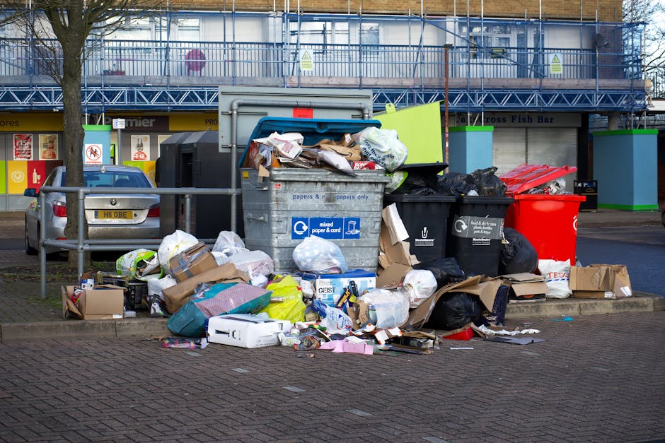 A large outdoor waste collection area on a paved urban street, featuring multiple overflowing rubbish bins and bags filled with various types of waste. In the foreground, there is a prominent grey mixed-paper-and-cardboard bin, its lid partially open with assorted newspapers, packaging, and cardboard boxes spilling out onto the surrounding pavement. Adjacent to this, black and black-and-white recycling bins are visibly overloaded, with black plastic bags and loose waste piled beside them. A bright red bin stands nearby, alongside several flattened cardboard boxes and smaller paper waste scattered across the ground. The scene is set against a background of commercial buildings with a blue scaffolding structure partially enclosing the upper level, and storefronts with glass windows displaying signage. A parked car with a visible license plate is positioned behind a metal railing, and the overall lighting suggests an overcast day, emphasizing the cluttered and unkempt appearance of the waste area. This visual context aligns with private waste collection and rubbish removal services in urban environments, highlighting the need for professional waste management solutions such as those offered by [COMPANY_NAME].