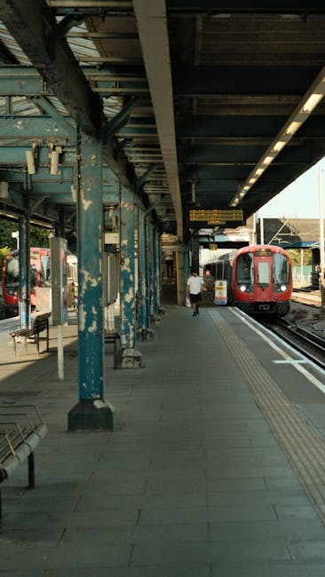 The image depicts a train station platform with a red commuter train arriving on the right side, partially visible as it stops at the station. The platform consists of grey paving slabs with tactile paving strips near the edge for accessibility. On the left, there are weathered blue metal supports and columns holding up an overhanging roof structure, which has a dark, industrial finish with some visible rust and peeling paint. A few benches are positioned along the platform, and a solitary person, dressed in casual clothing, is walking towards the train in the middle distance. The background shows additional platforms and tracks, along with some advertising boards overhead. The lighting indicates daylight, with the shadows highlighting the structural details of the platform's roofing and supporting framework, typical of an urban railway station where independent or alternative rail services may operate. This scene exemplifies a typical public transport setting, where the station infrastructure supports passenger movement and train arrivals, with a focus on safety features like tactile paving and clear signage for efficient boarding and alighting, consistent with local railway standards for passenger convenience and safety.