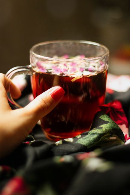 A close-up image of a clear glass mug filled with herbal tea, which has a dark amber colour and floating dried flower petals on the surface. The mug is being held by a person's left hand, with neatly manicured nails painted red, positioned on the left side of the image. The mug rests on a dark, floral-patterned fabric with red, pink, and green hues, likely part of a blanket or clothing item. The background is blurred and indistinct, focusing solely on the tea and hand, creating a warm, intimate atmosphere. This image subtly relates to the context of private or alternative waste handling by emphasizing a domestic setting, free from large waste or rubbish containers, aligning with a focus on home-based rubbish removal or disposal practices, such as those managed by independent rubbish collection services like RubbishCollectionEarlscourt.co.uk.
