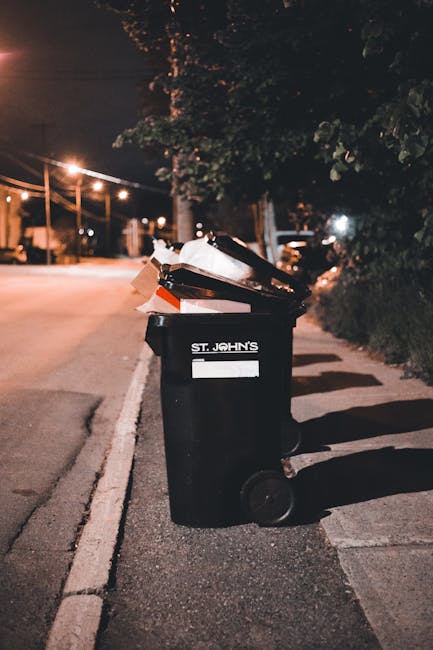 A black wheeled rubbish bin marked with the label 'St. John’s' is positioned on a sidewalk at night, partially filled with discarded cardboard boxes, paper, and other waste materials. The bin's lid is slightly open, revealing the mixed waste inside. The surrounding environment includes a dark pavement border, a street illuminated by warm orange streetlights, and trees with dense foliage visible in the background, casting shadows over the pavement. The scene suggests an instance of private waste disposal or on-site rubbish management outside residential or commercial premises, possibly in an area serviced by independent collection providers like St. John’s rubbish removal. The overall atmosphere is quiet, with no movement or other activity visible, highlighting the bin's role in waste management for local properties, aligning with services typical of rubbish collection and private disposal solutions in urban settings.