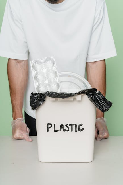 A black wheeled rubbish bin marked with the label 'St. John’s' is positioned on a sidewalk at night, partially filled with discarded cardboard boxes, paper, and other waste materials. The bin's lid is slightly open, revealing the mixed waste inside. The surrounding environment includes a dark pavement border, a street illuminated by warm orange streetlights, and trees with dense foliage visible in the background, casting shadows over the pavement. The scene suggests an instance of private waste disposal or on-site rubbish management outside residential or commercial premises, possibly in an area serviced by independent collection providers like St. John’s rubbish removal. The overall atmosphere is quiet, with no movement or other activity visible, highlighting the bin's role in waste management for local properties, aligning with services typical of rubbish collection and private disposal solutions in urban settings.
